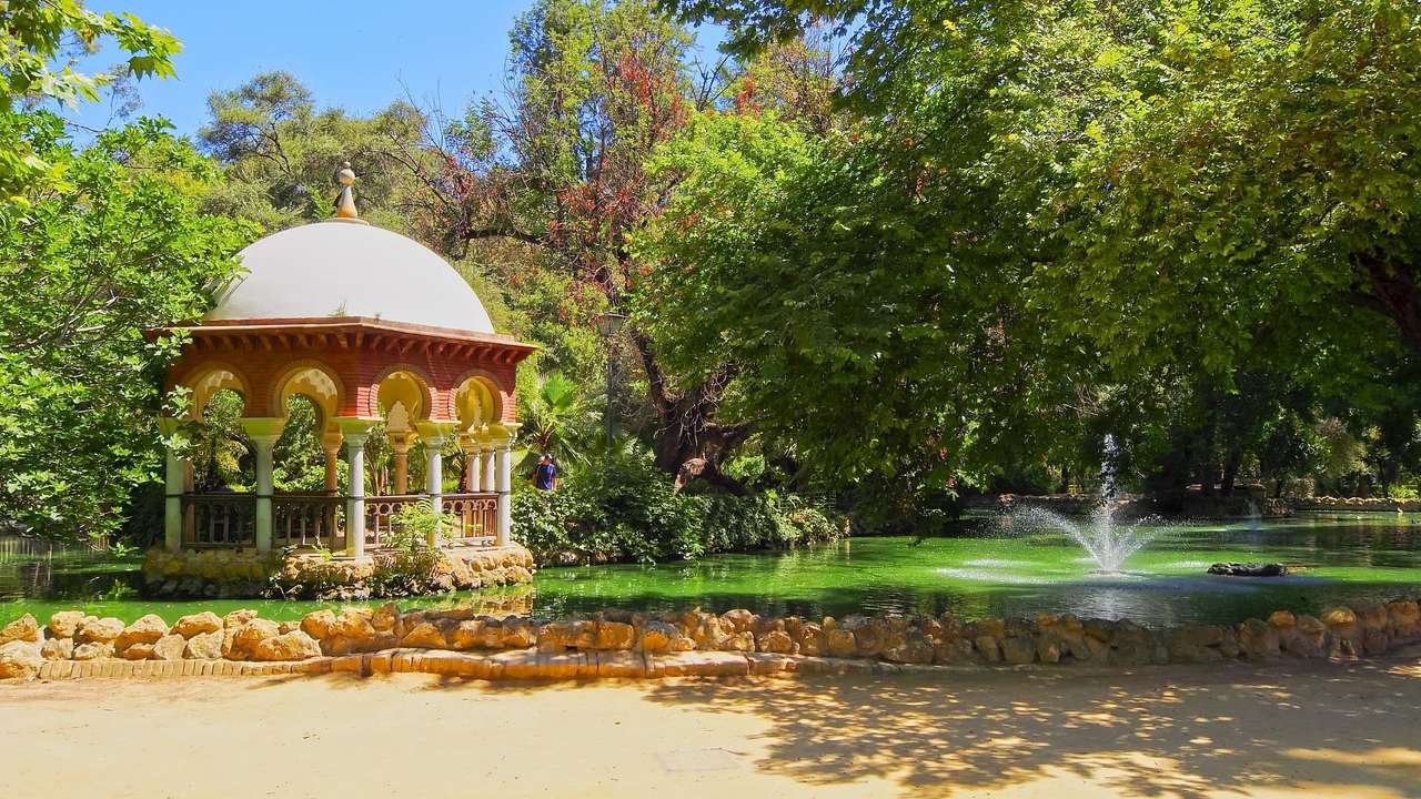 A small structure with arches and a domed roof in a park with trees and a fountain