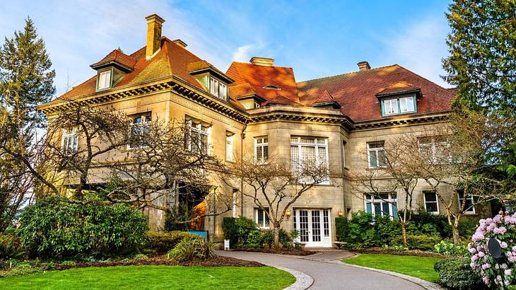 A brown mansion with a red roof, surrounded by trees & grass under a blue sky