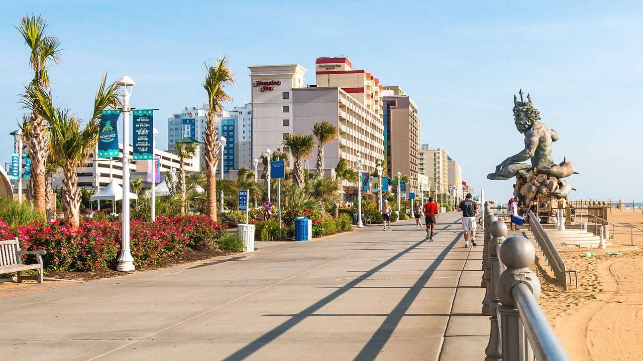 A beachside boardwalk surrounded by palm trees, buildings, sand, and a Neptune statue