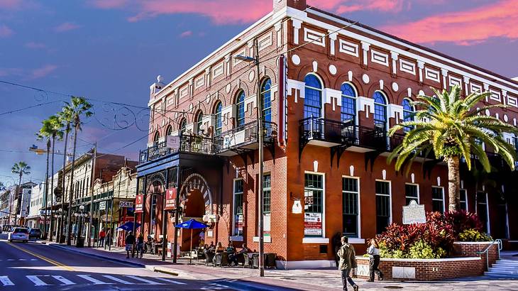 A red brick building, palm trees, and a street under a pink and purple sky