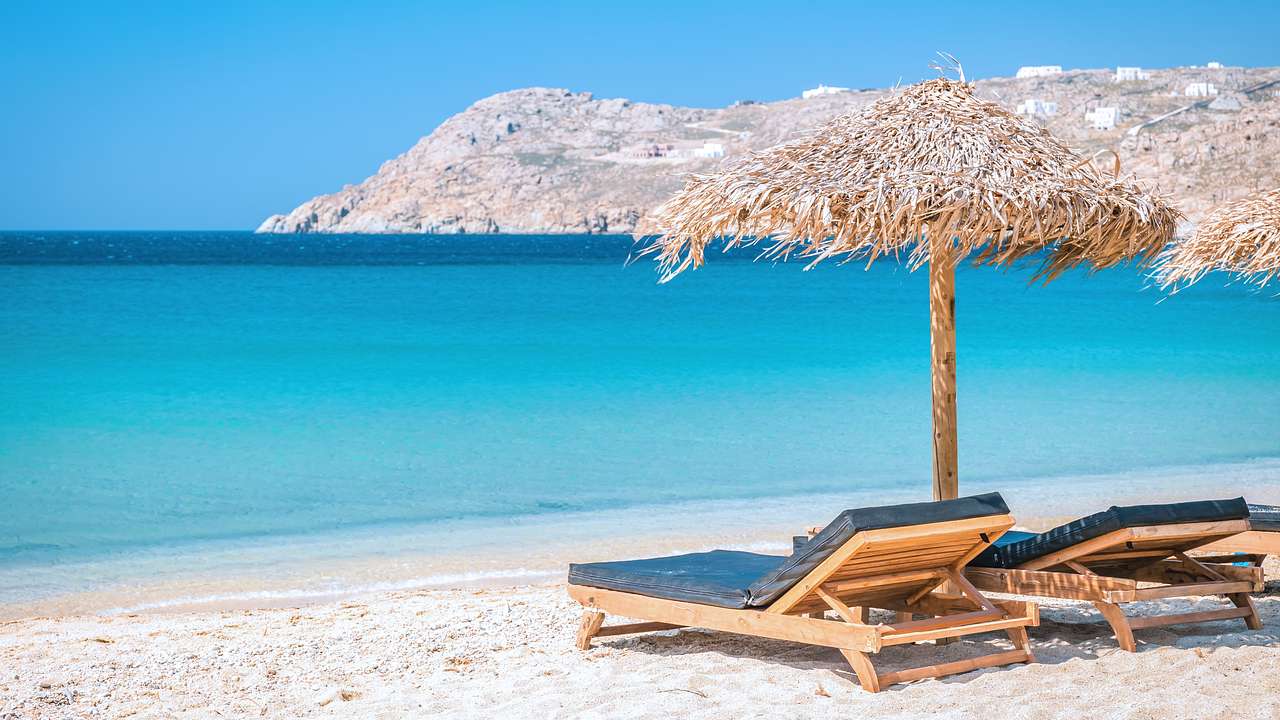 Lounge chairs under a sun umbrella by a sandy beach and the bright blue ocean