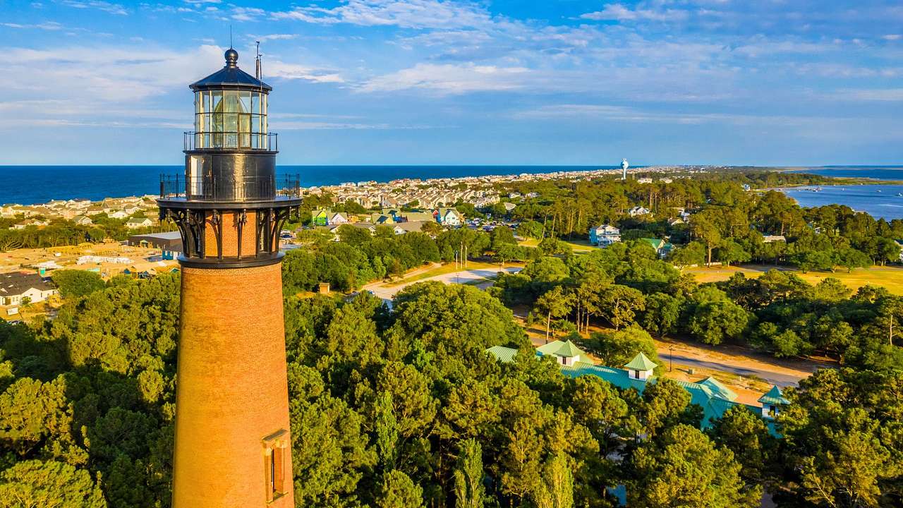 A tall brick lighthouse surrounded by trees under a blue sky with clouds