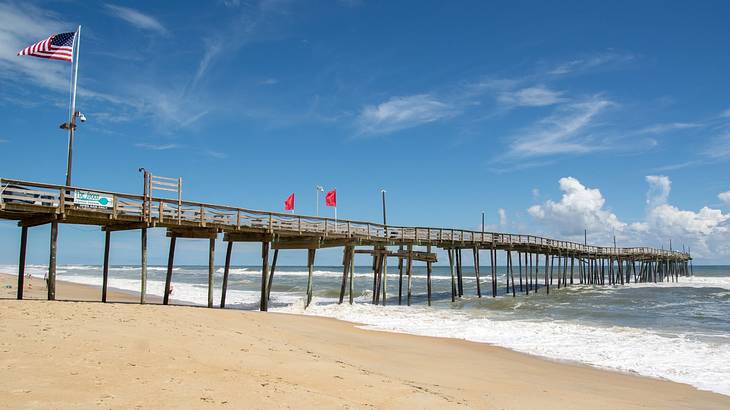 A long wooden bridge extending from the shore to the sea with multiple flags