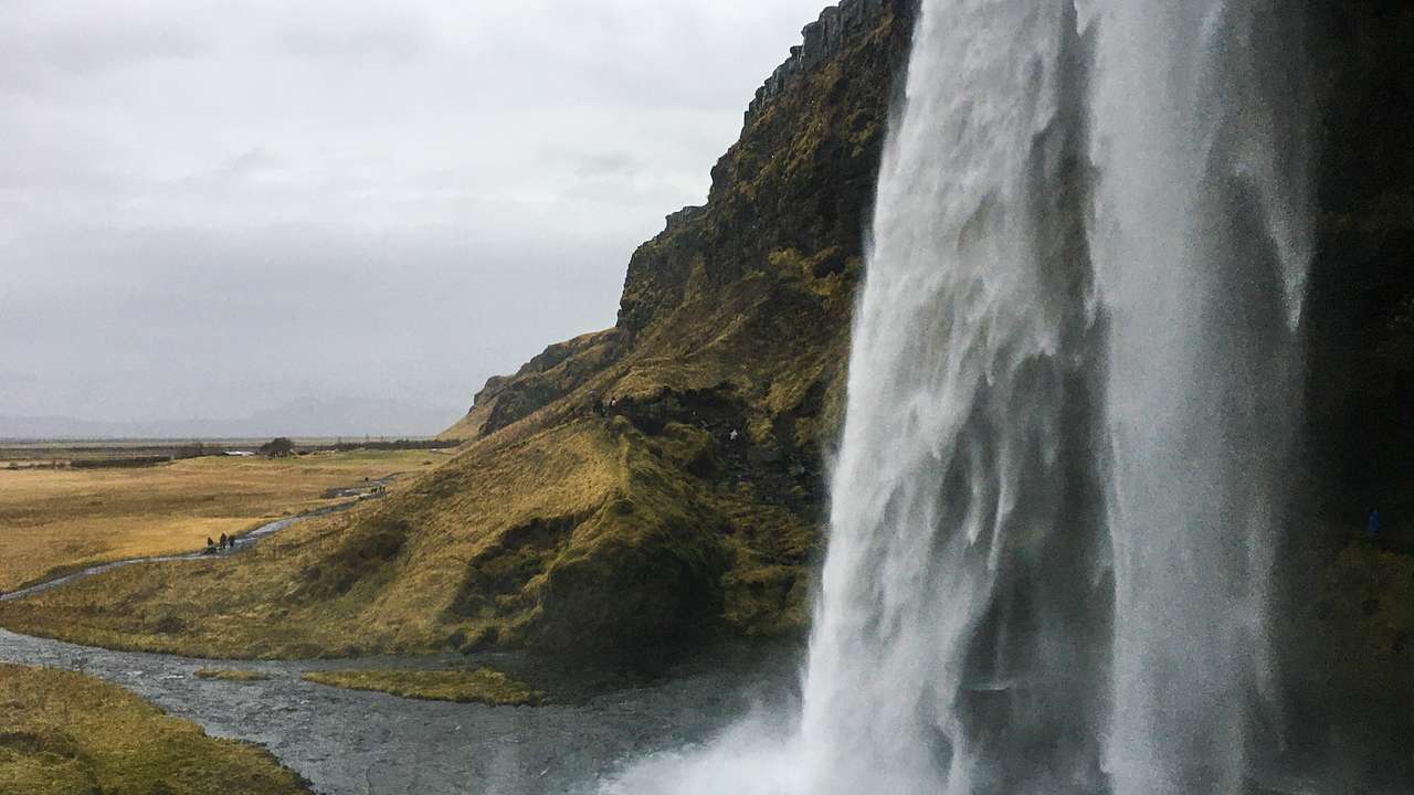 A waterfall flowing into a pool next to greenery under a cloudy sky