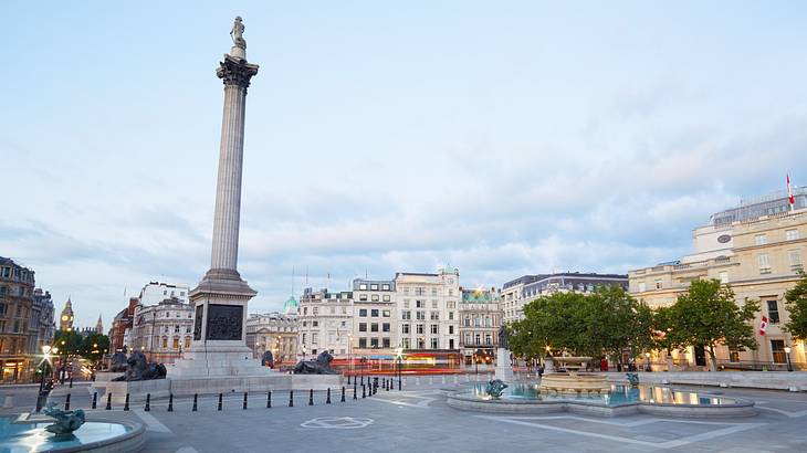 A column with a statue atop it in a square with a fountain and buildings