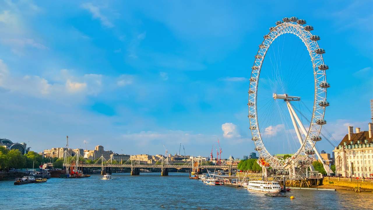 A Ferris wheel next to buildings and a river with a bridge over it under a blue sky
