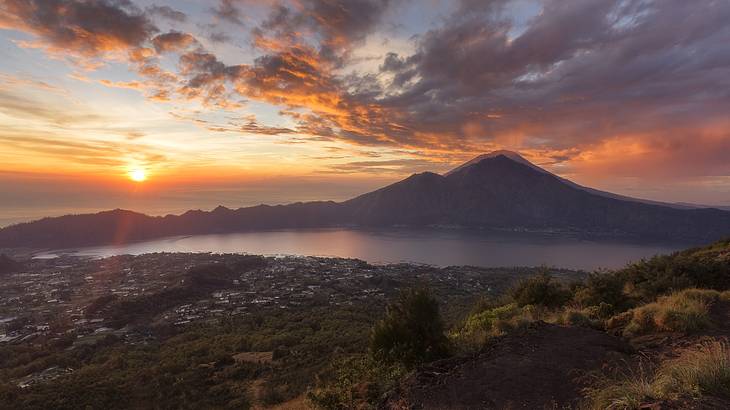 Greenery next to a lake and mountains under an orange sky with clouds at sunrise