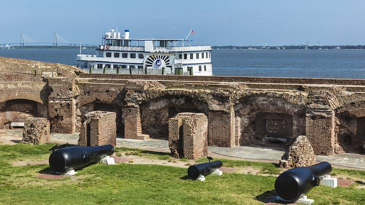 Three black cannons by a brick wall of a fort with a ship on the sea in the back