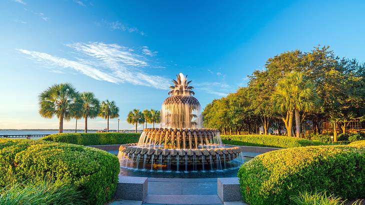 A pineapple-shaped fountain surrounded by greenery in a park by the water