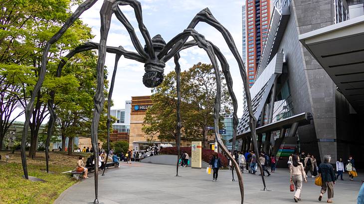 A large spider sculpture on a path walk surrounded by buildings, trees and people