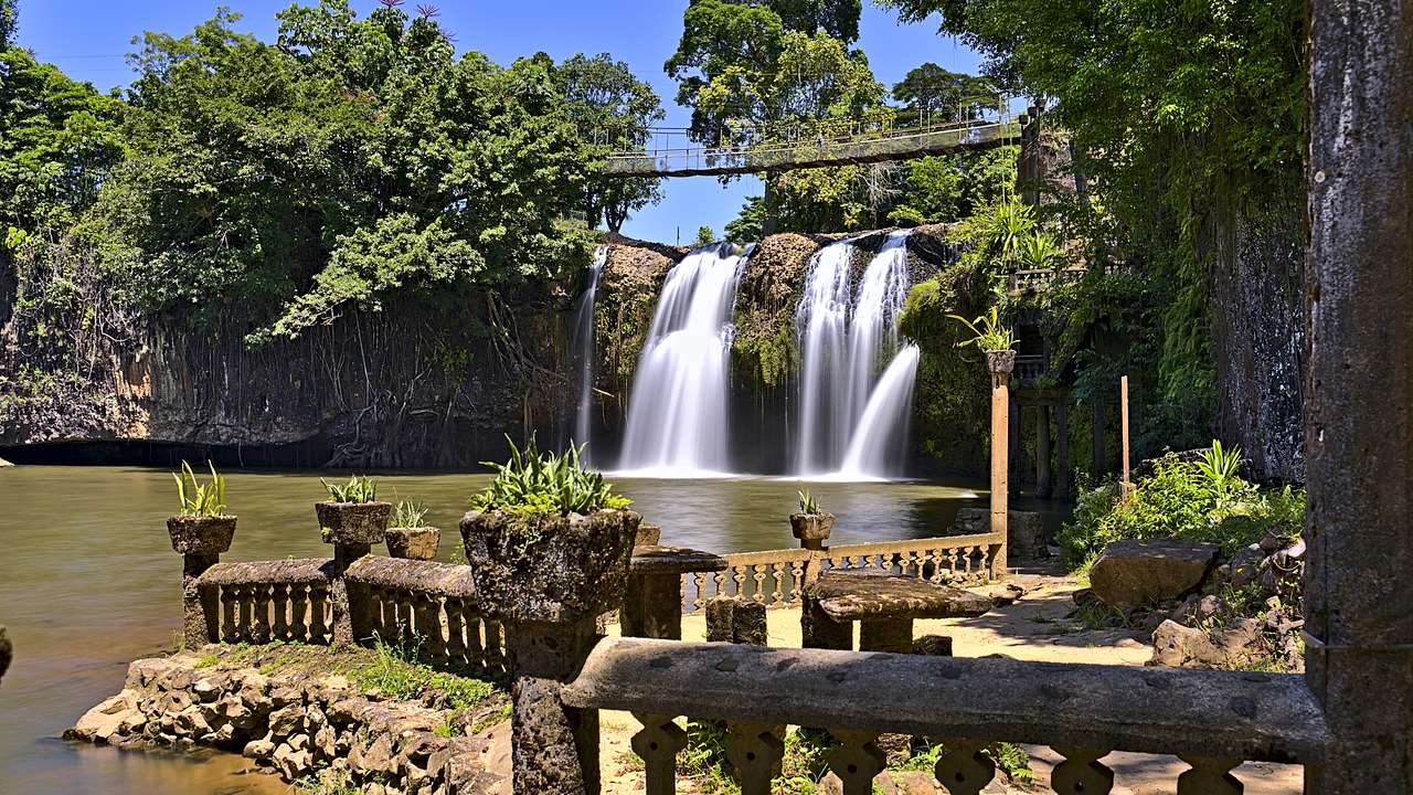A waterfall surrounded by trees facing a bench and porch area on a sunny day