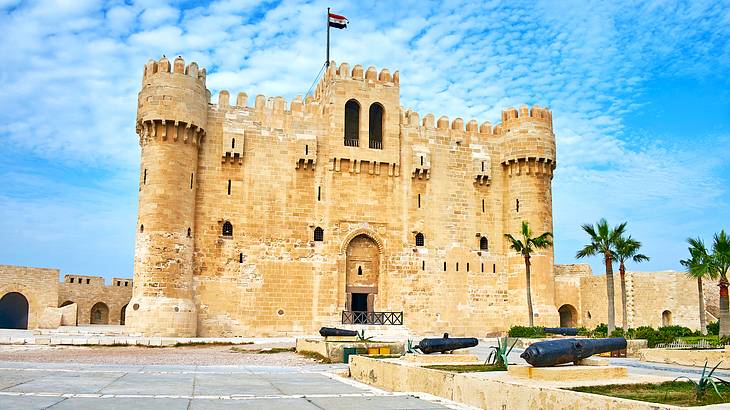 The medieval fort Citadel of Qaitbay with a flag on top facing trees and cannons