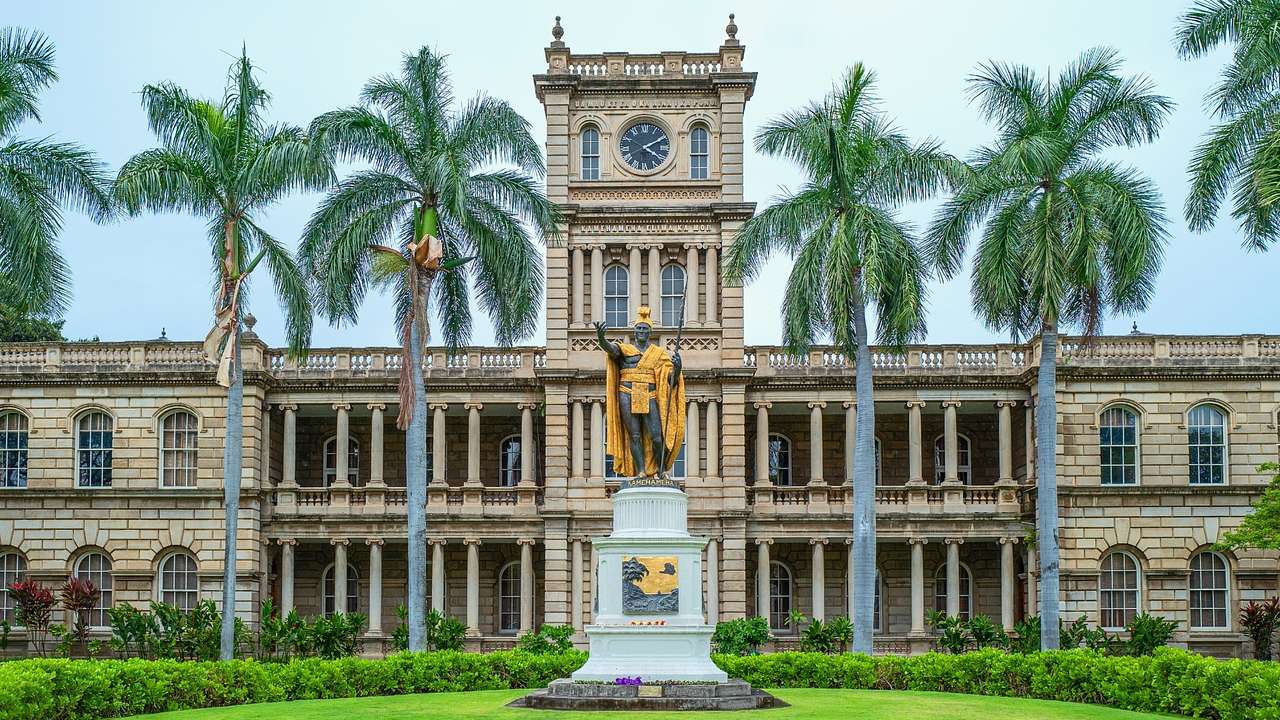 A gold-covered statue in front of a palace-style building with grass and palm trees