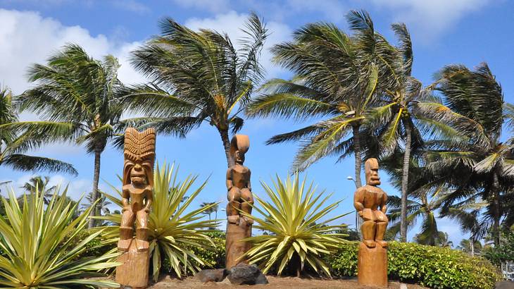 Three Polynesian statues on grass with palm trees and blue sky behind them