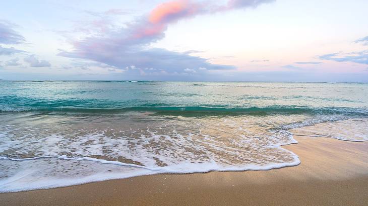 A soft sand beach with the water rushing to the shore underneath a partly cloudy sky