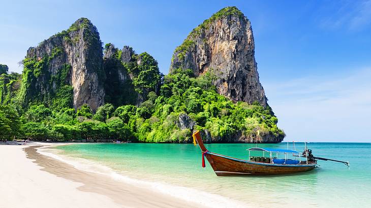 A long-tail boat by the beach near tall cliffs with greenery on them