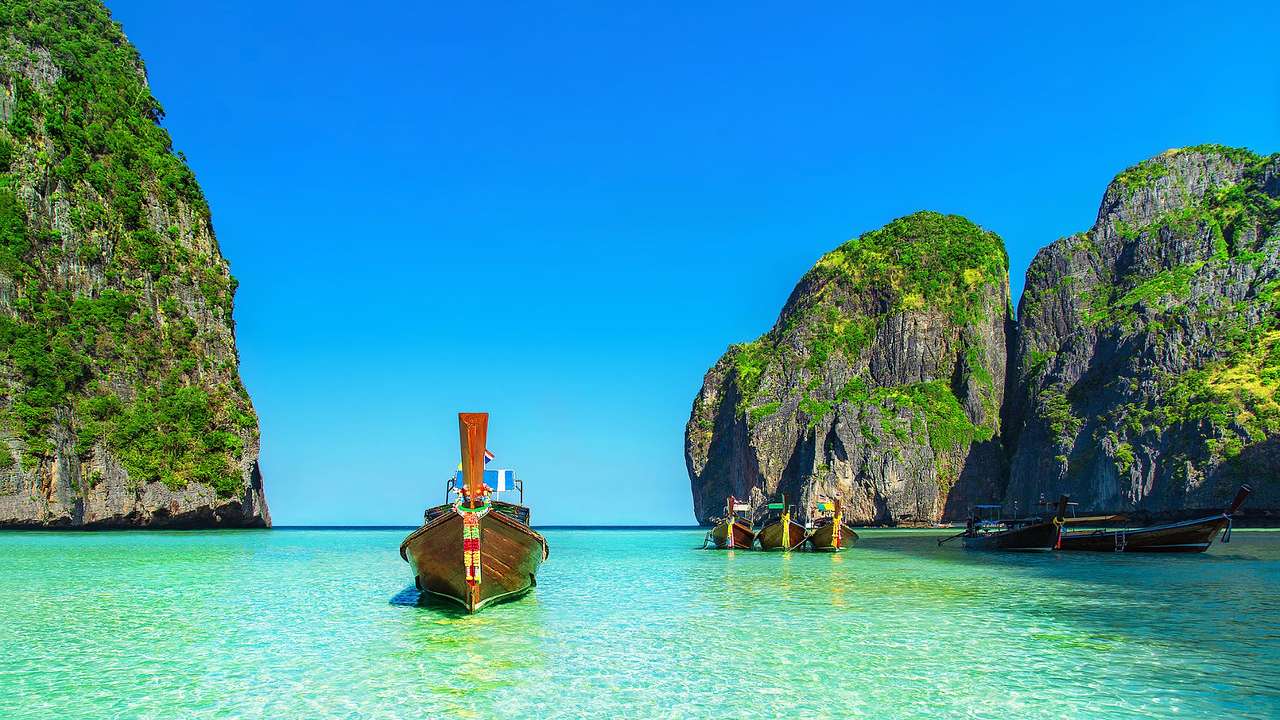 A long-tail boat on blue water next to greenery-covered cliffs and a blue sky
