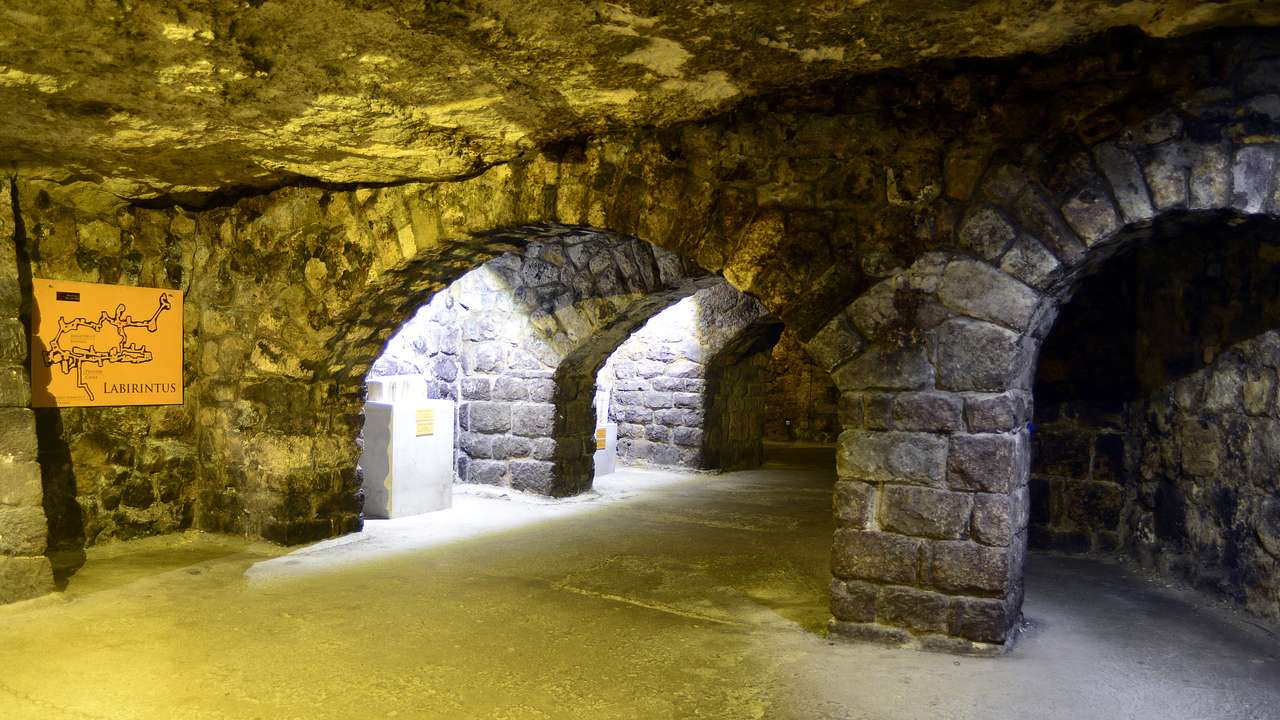 A low-ceiling stone hallway with arched doorways and a map of a labyrinth on the wall