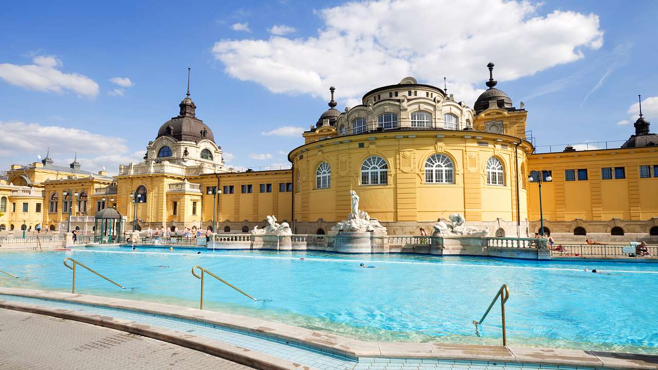 A large pool with some statues on the side and a large yellow building on a nice day