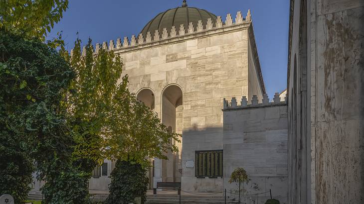 A white stone building with a dome, arched entryways, and trees in front