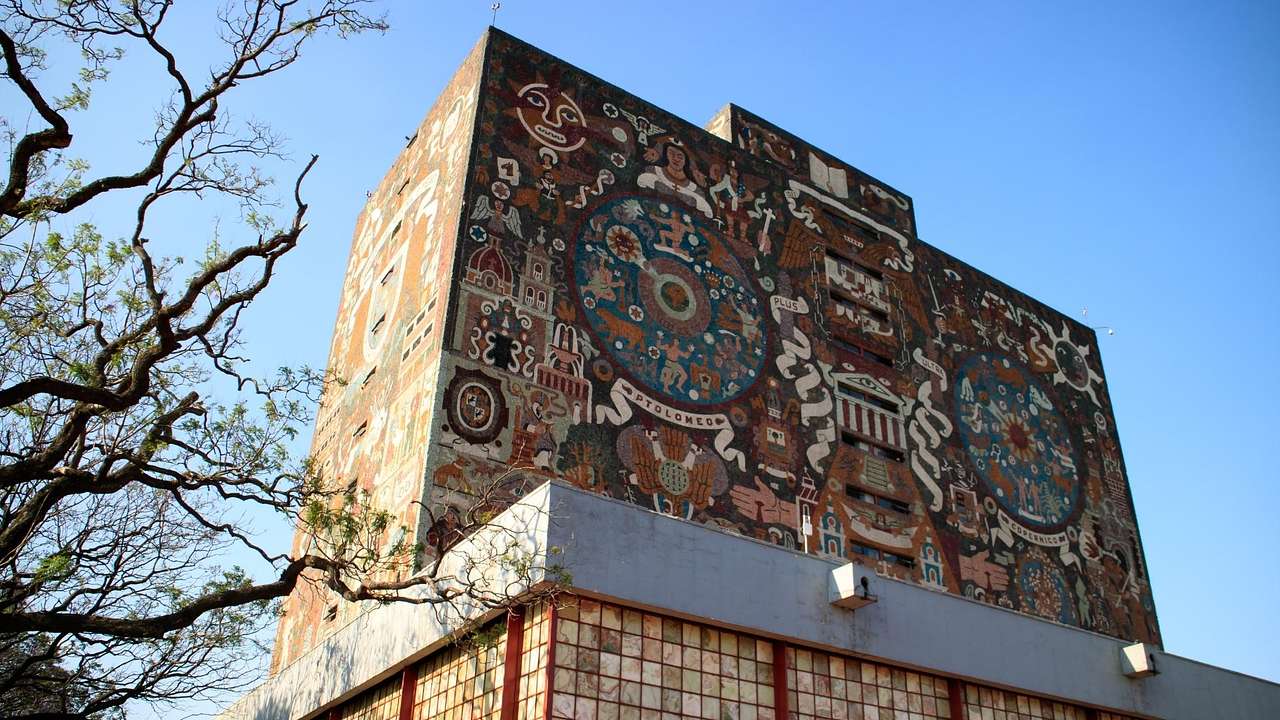 Looking up at a brown building filled with mosaic-tiled murals against a clear sky