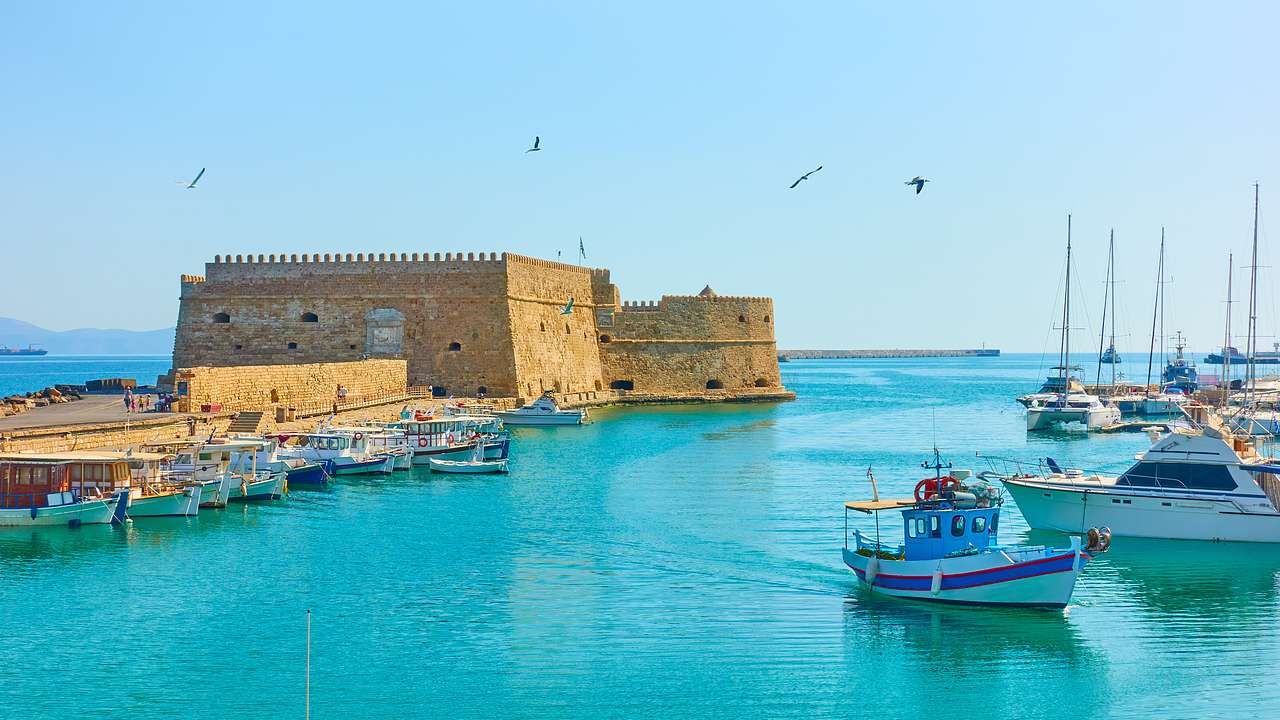 An old bricked fortress near the sea with docked ships and birds flying over