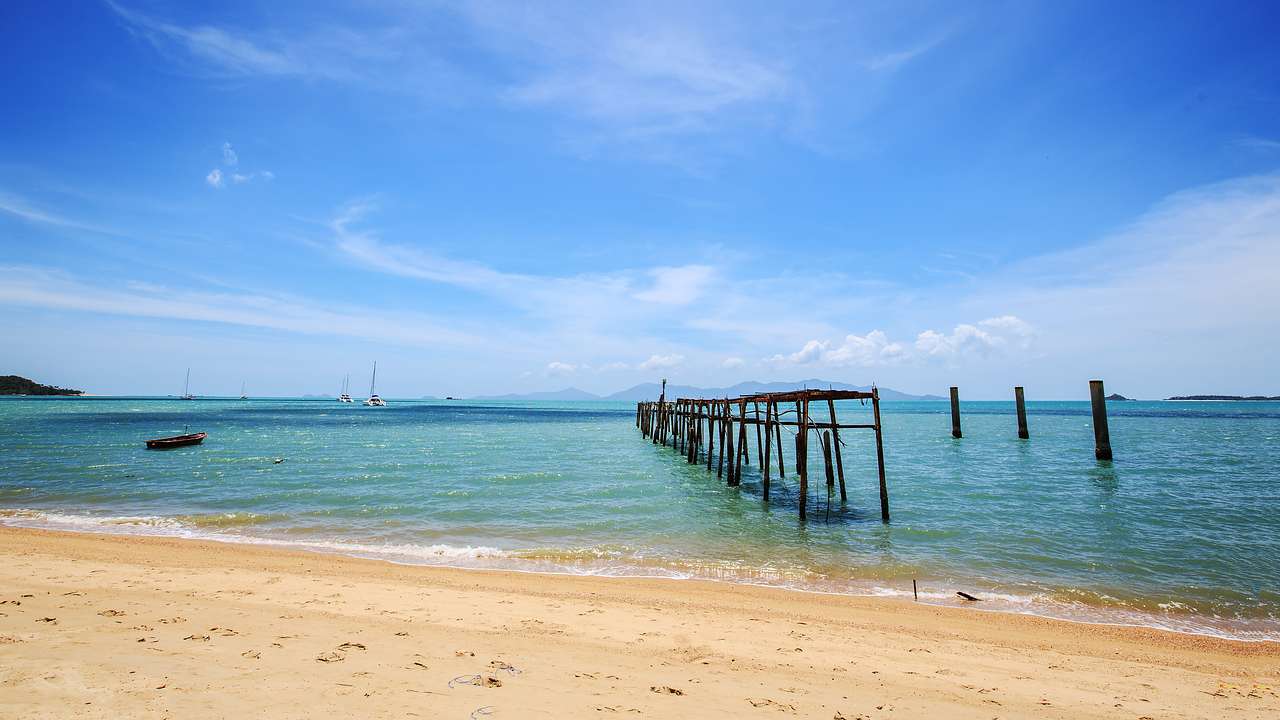 An old wooden pier in the water near a sandy shore under a blue sky with some clouds