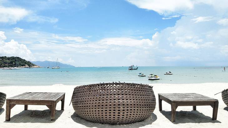 Wooden tables and a rattan lounge chair near the beach with boats in the background