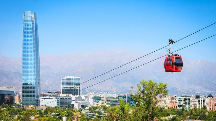 A red cable car over trees with buildings, a skyscraper, and mountains next to it
