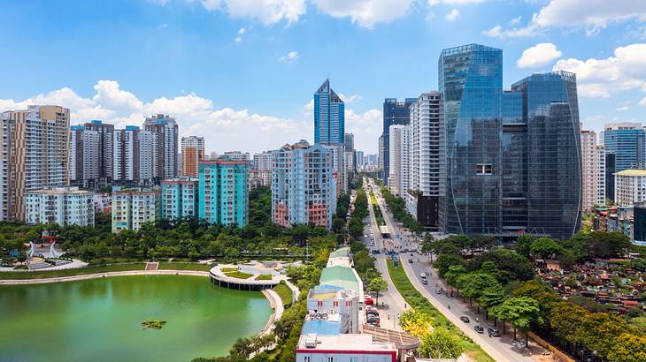 A city skyline next to a road, a lake, and green trees under a blue sky with clouds