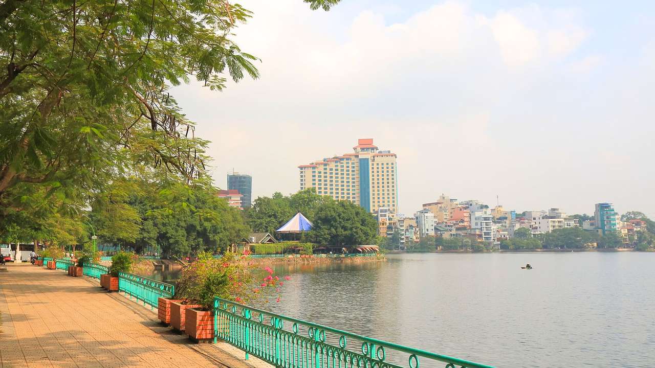A fenced walkway next to the water, a tall building, and other smaller buildings