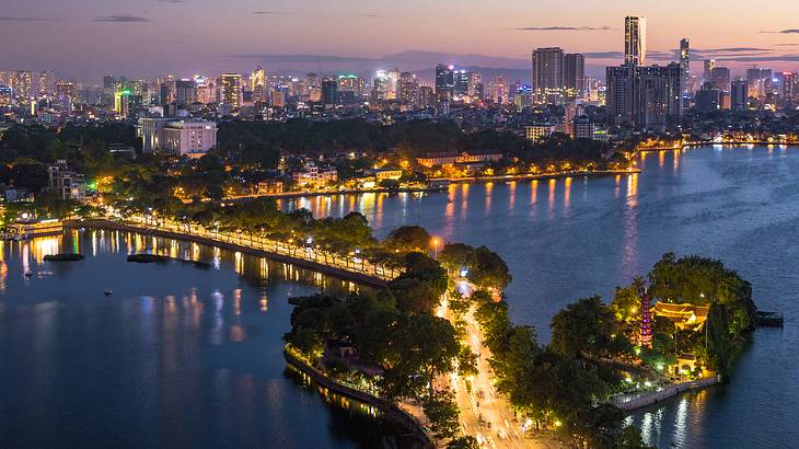 Aerial view of a city skyline at night, next to the water and a road with trees