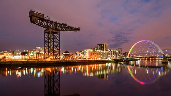 A crane and an illuminated arch next to a river and lit up city buildings at night