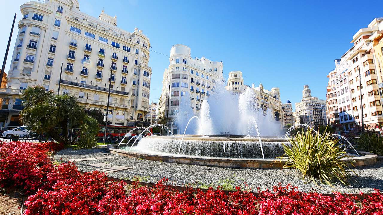A fountain surrounded by red flowers and tall white buildings