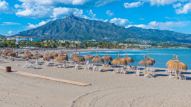 Straw umbrellas and lounge chairs by the beach with a mountain in the background