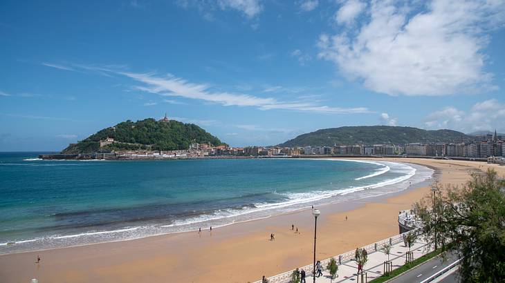 A sandy beach next to the ocean, a greenery-covered hill, and a promenade