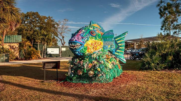 A sculpture of a green fish made with trash on grass against trees & parked cars