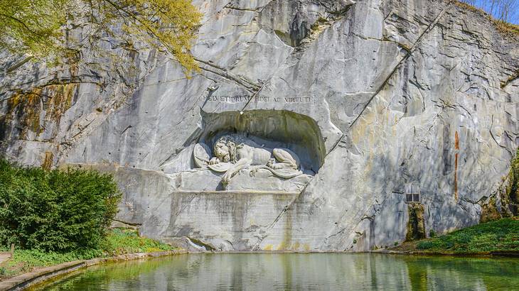 A lion carved into a rock face next to a pond and trees