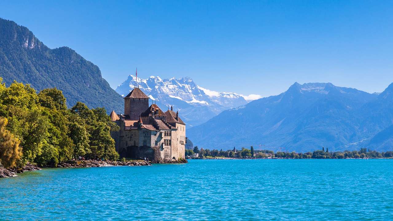 A vast lake next to trees, a castle, and mountains on a clear day