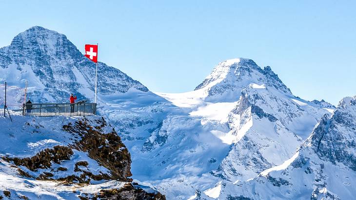 Snow-covered mountains next to a viewing platform with a swiss flag and people on it