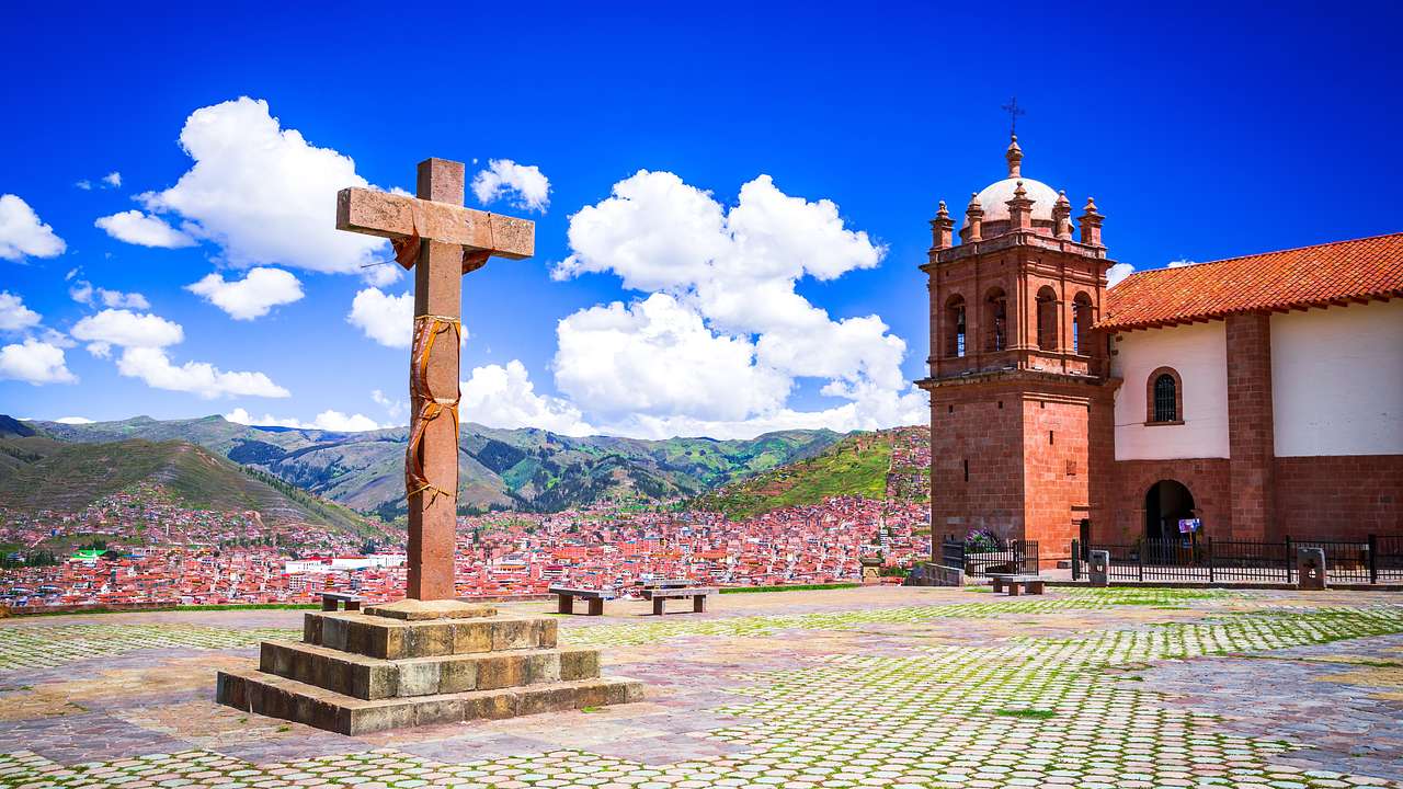 A large crucifix in the middle of a tiled open area near a church with a bell tower