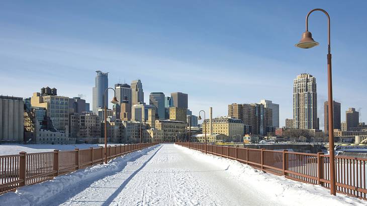 A snow-covered path on a bridge with an icy river underneath and buildings in front