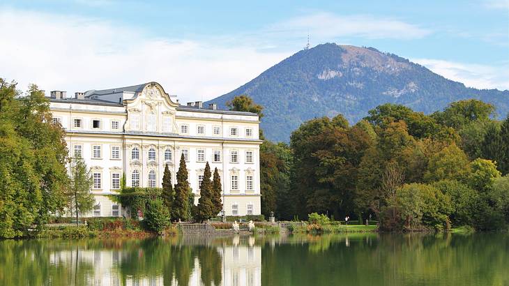 A large white manor house near a lake and trees with a mountain in the background