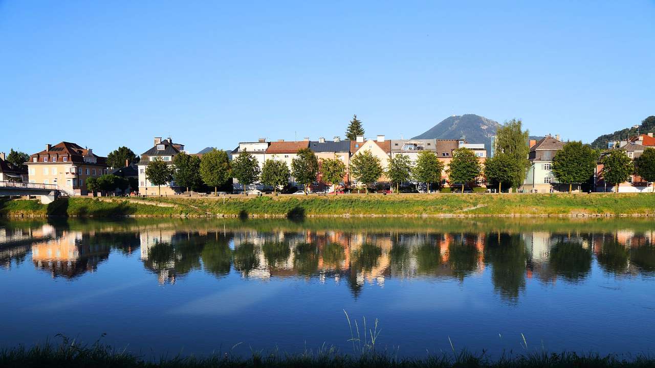 A lake next to houses, trees, and a greenery-covered hill under a blue sky