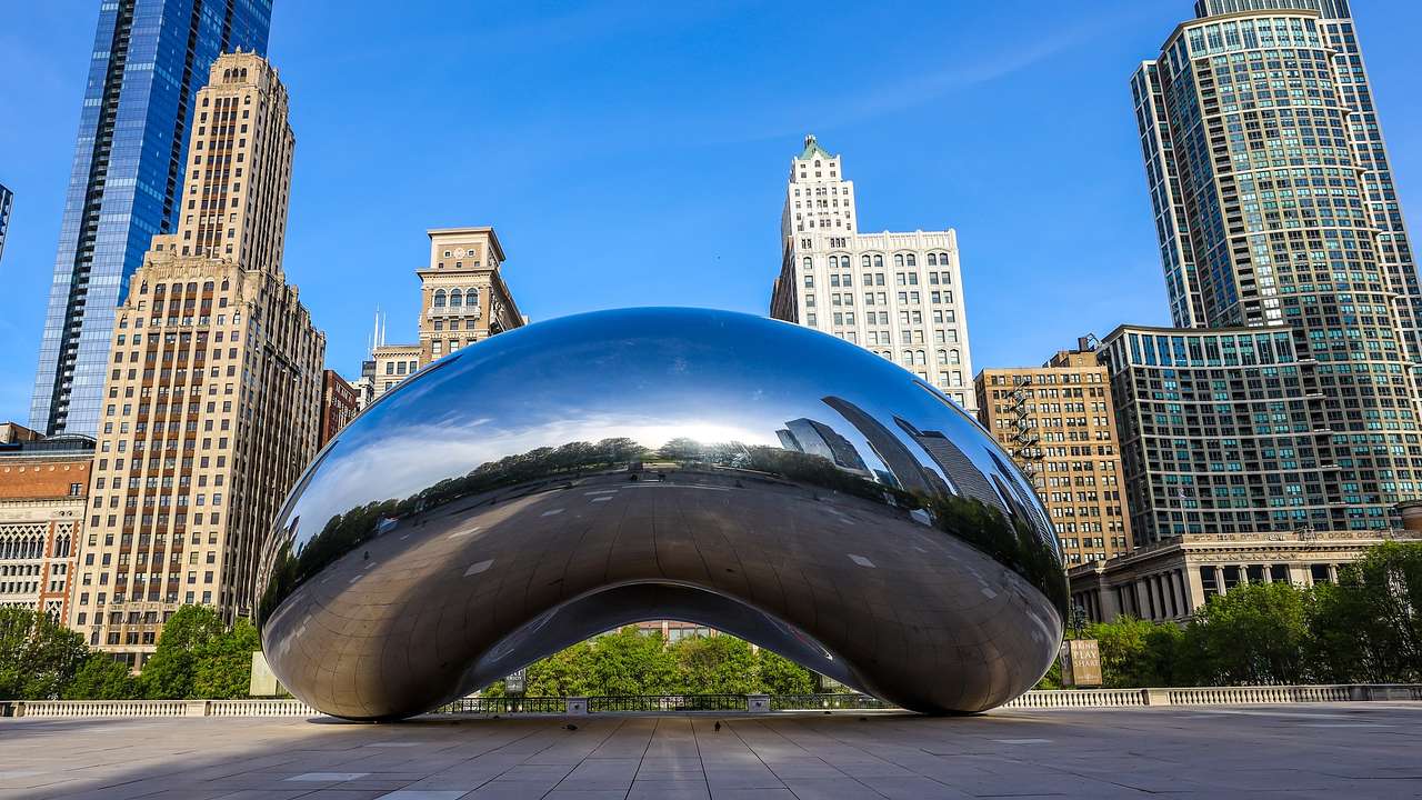 A large bean-shaped metallic structure on a walkway with high-rise buildings behind