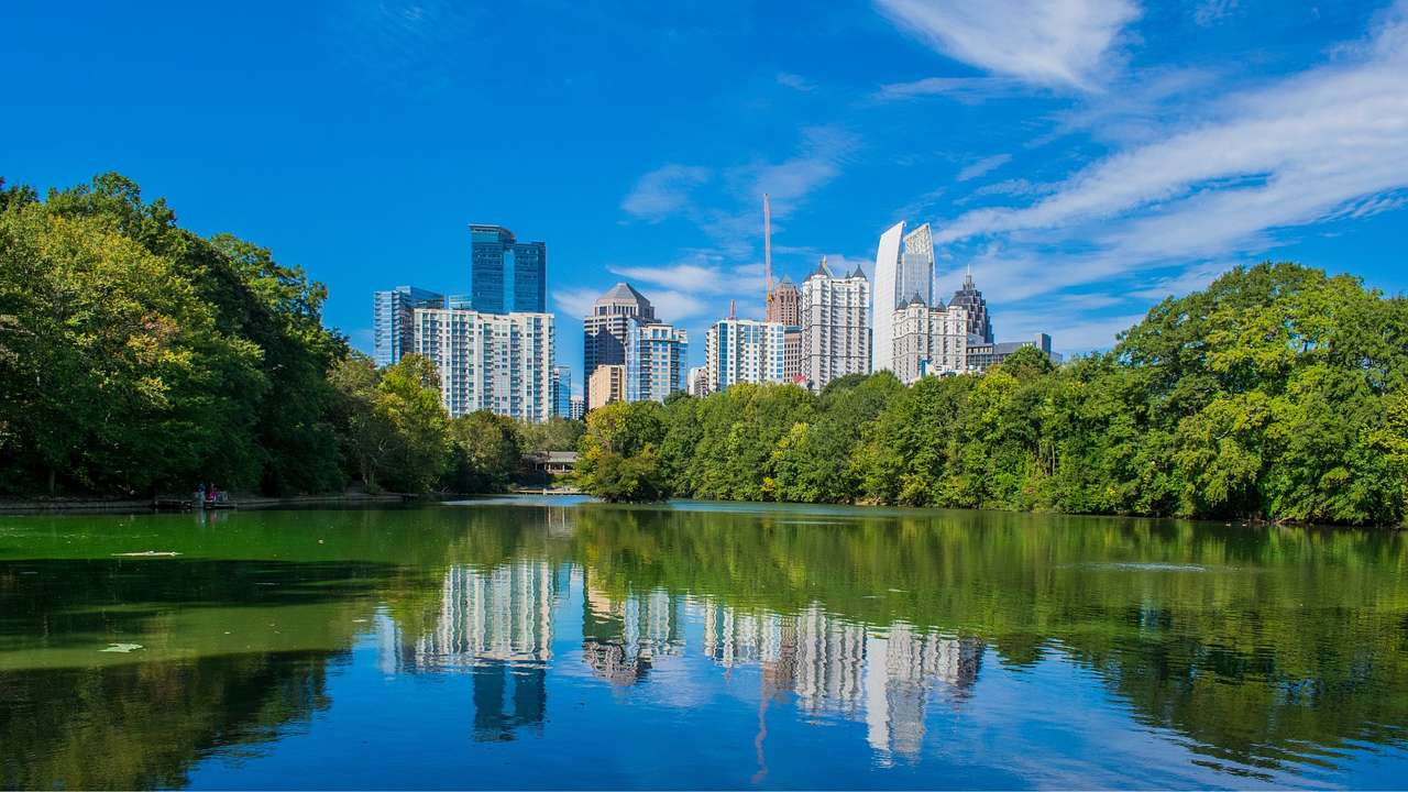 A lake surrounded by trees with a city skyline behind them under a blue sky