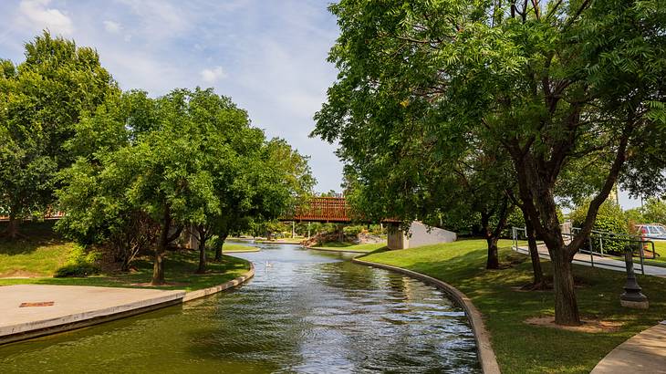 A park with trees, a paved walkway, and a bridge over a canal running through it