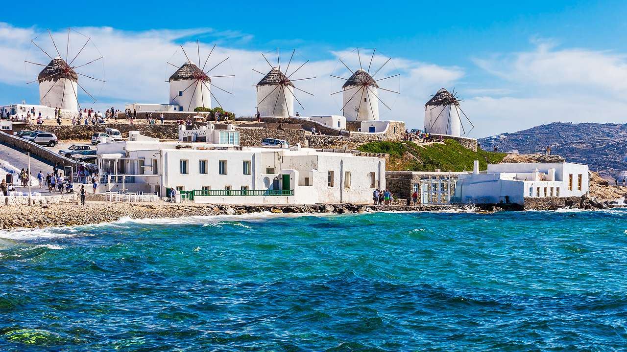 Windmills on a green hill with water and rocks in the foreground, Mykonos, Greece