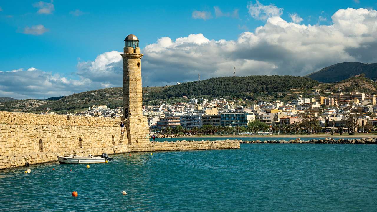 A stone lighthouse structure next to the water and a city with small houses by a hill