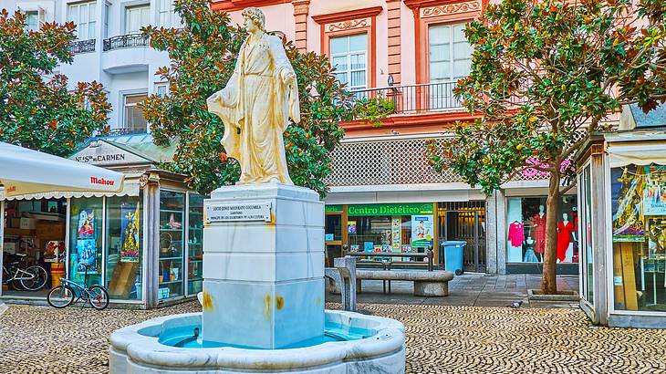 A marble fountain with a human statue on top surrounded by shops and trees